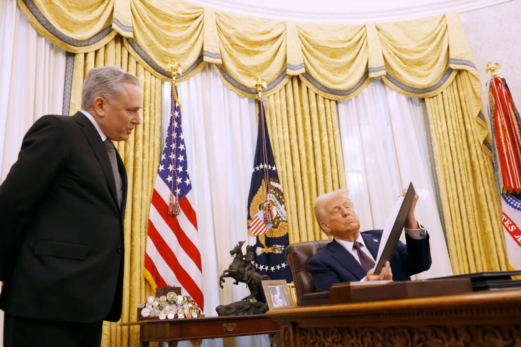 Donald Trump reviewing a framed document at the Resolute Desk with US flags visible in the backgroun