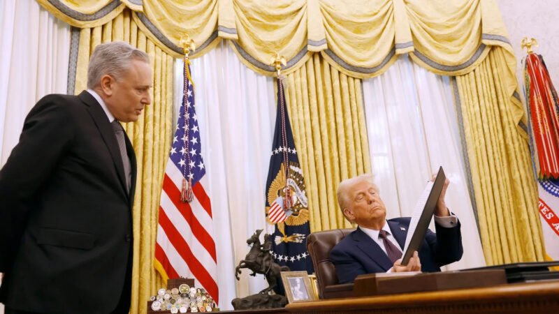 Donald Trump reviewing a framed document at the Resolute Desk with US flags visible in the backgroun
