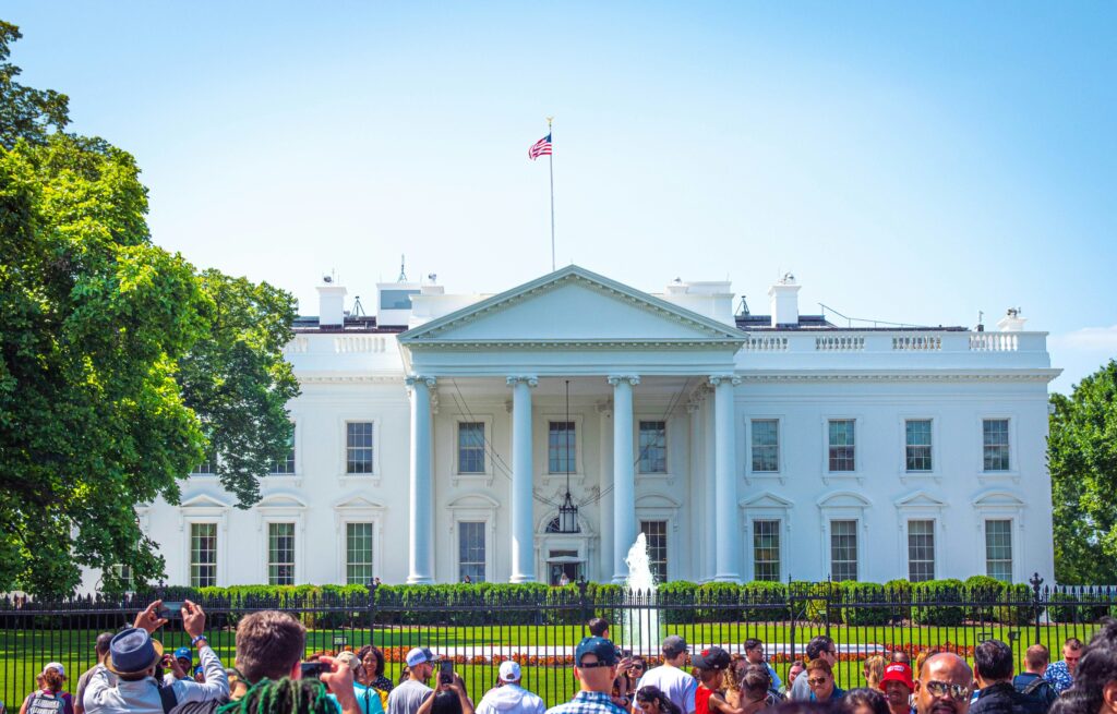 White House building in Washington, DC with the US flag flying above, surrounded by trees, a fountain, and visitors outside.